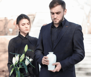 A man holds an urn and a woman stands next to him as he appears to be addressing a memorial service.