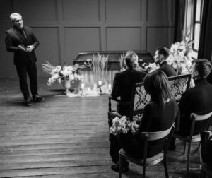 A man stands in front of a group of mourners at a memorial service. Next to him is the casket decorated with candles.