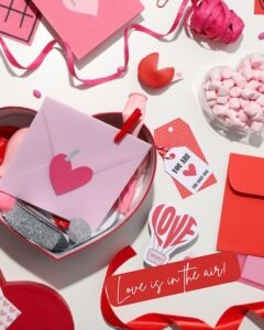 A table covered in Valentines Day items, candy, bows, letters and paper hearts. Love is in the air reads at the bottom.