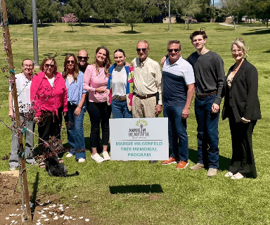 A group gathers around a tree planting including a sign that reads The Margie Hilgenfeld Tree Memorial program.
