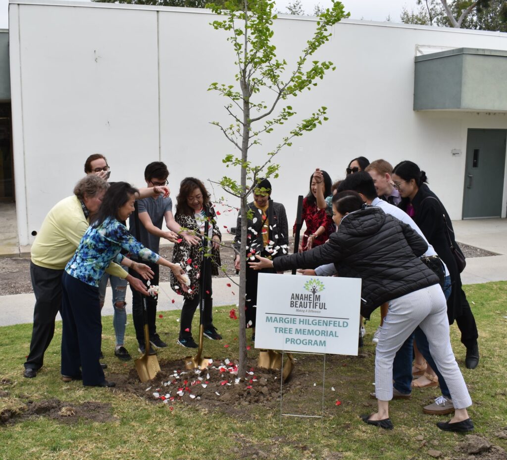 Family members gather around a tree which has been planted in honor of a loved one. Rose petals are scattered at the trunk.