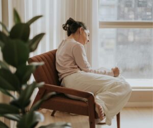 A young woman curled up in a chair writing something in front of a lighted window.